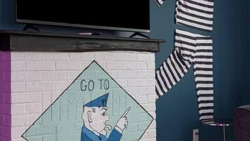 Una habitación con un televisor montado en la pared, un conjunto de pijama a rayas colgado en la pared, y un mural con un personaje señalando 'JAIL'.