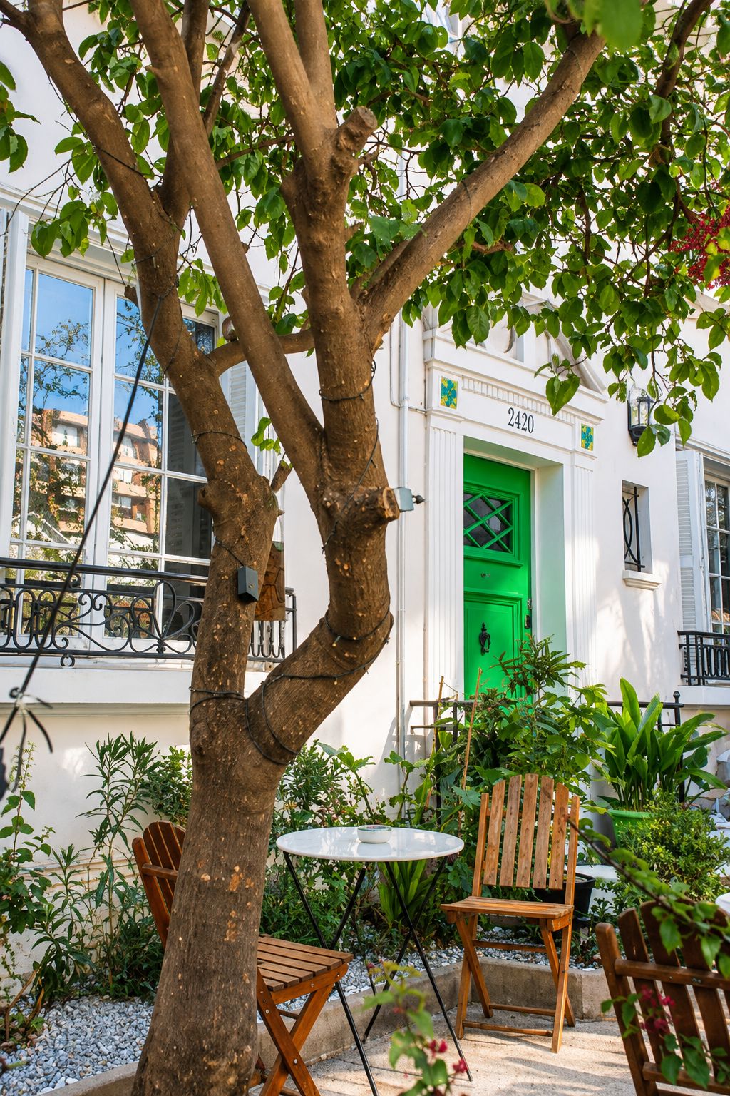 Un espacio al aire libre con bancos bordeados de árboles, una puerta verde, un edificio blanco con el número 2420 y un balcón con barandilla.