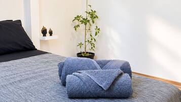 Una cama bien tendida con una manta azul, una planta en maceta y una almohada negra sobre una pared blanca.