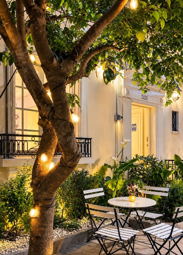 Un espace extérieur aménagé avec des sièges, un arbre, des guirlandes lumineuses et l’entrée d’un bâtiment.
