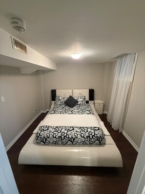 A bedroom with a bed, a bedside table, a nightstand, a ceiling vent, and a window with curtains. - One bedroom basement apartment (Vaughan)