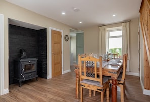 A dining area with a wooden table and chairs, a black wood-burning stove, and a window with curtains. - Newly Renovated Farmhouse (KY)