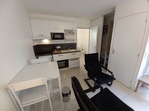 A compact kitchenette with a microwave, sink, and cabinets. There is a dining table with chairs and a small stool. A black office chair is also present. - 4-Star Furnished Studio (West-Facing) (JONZAC)