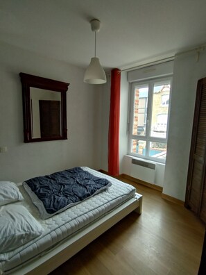 Ein Schlafzimmer mit einem Bett, einer Pendelleuchte, einem Fenster mit Blick auf Gebäude und einer Heizung.