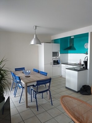 Eine moderne Küche mit weißen Schränken, einer Türkis-farbenen Akzentwand, einem Essbereich mit einem Holztisch und blauen Stühlen sowie einer hängenden Pendelleuchte.