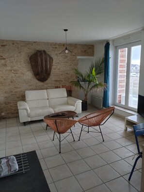 Ein Wohnzimmer mit Steinsandsteinwand, einem weißen Sofa, einem Couchtisch, zwei Korbstühlen, einer Topfpflanze und einem großen Fenster mit Vorhängen.