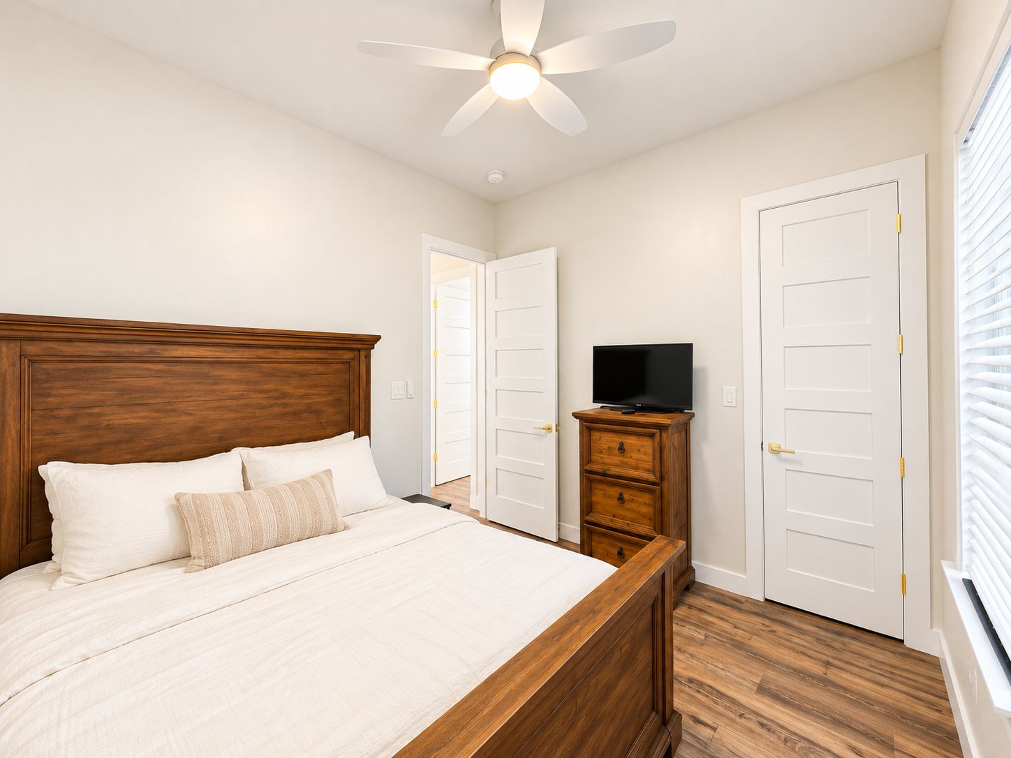 A bedroom with a wooden bed, a ceiling fan, a television on a wooden dresser, and white doors.