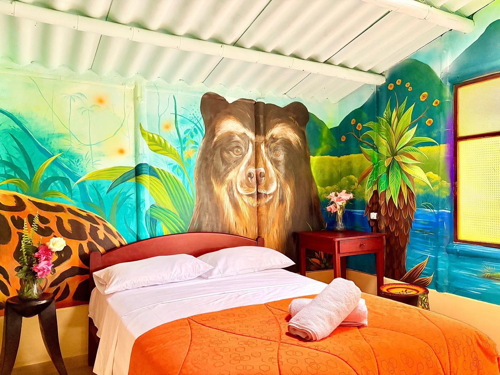 A room with a bed, a large bear mural, a vase with flowers, and a view of a blue body of water.