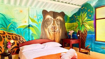 A room with a bed, a large bear mural, a vase with flowers, and a view of a blue body of water.