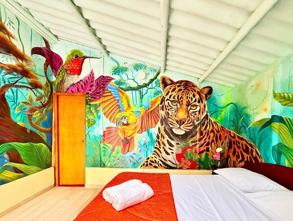 A room with a mural of a tiger and a parrot, a wooden cabinet, and a bed with a red and white patterned bedspread.