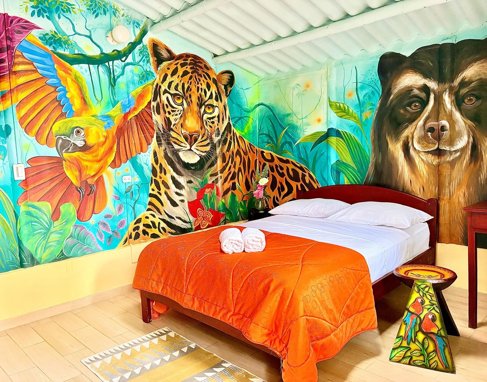Un dormitorio con un mural que muestra un tigre, un loro y un león. Hay una cama con una colcha naranja y una mesita con un diseño de loro.