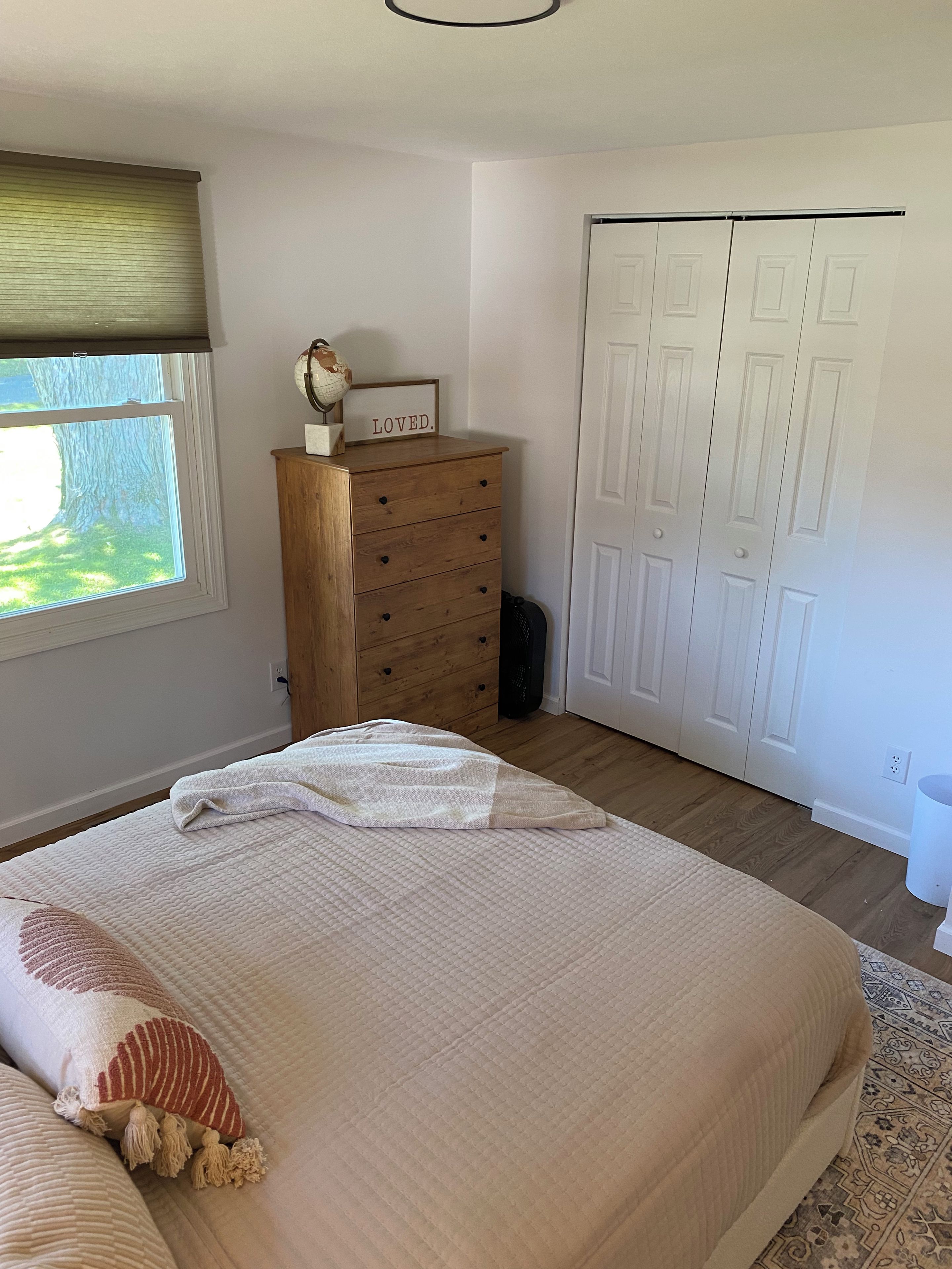 A bedroom with a bed, a wooden dresser, a window with blinds, a white door, and a rug.