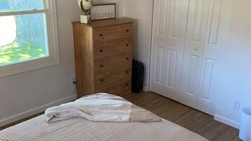 A bedroom with a bed, a wooden dresser, a window with blinds, a white door, and a rug.