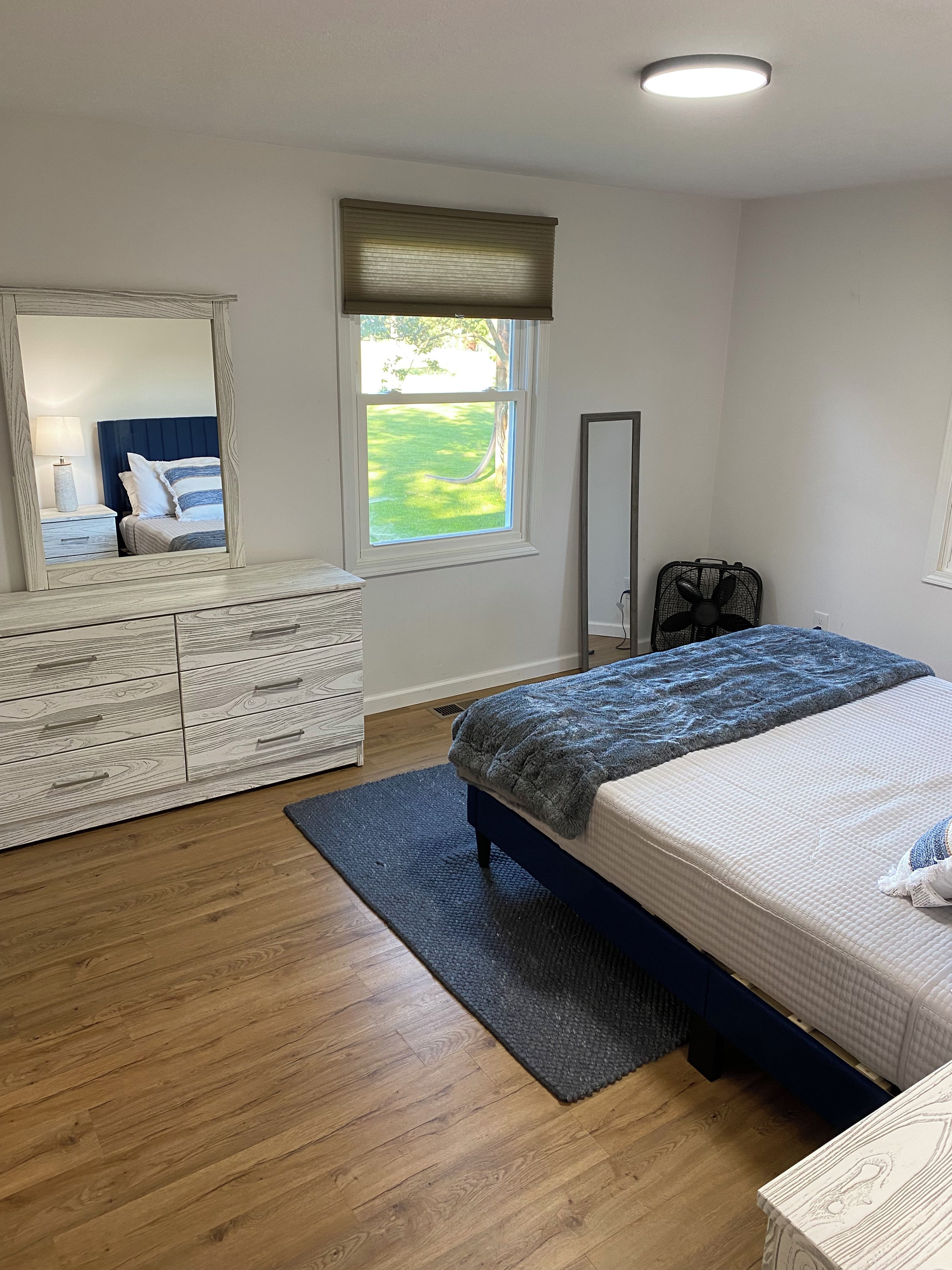 Une chambre à coucher avec un lit, une commode, un miroir, une fenêtre et un ventilateur.