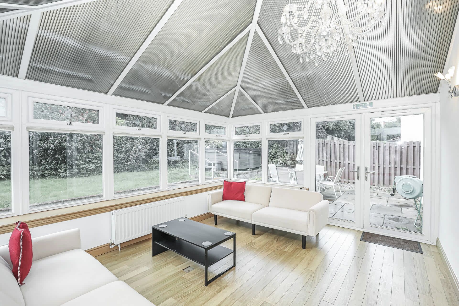 A conservatory with a white interior, large windows, a black coffee table, and a white sofa with red cushions.