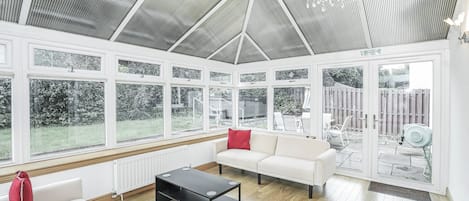 A conservatory with a white interior, large windows, a black coffee table, and a white sofa with red cushions.