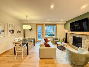 A modern living room with a fireplace, a dining area, and a view of the city through large windows. - Mountain Lynx Retreat | Spring Creek King Suite (Canmore)