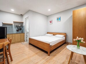 A bedroom with a wooden bed, a small table with a vase of flowers, and a kitchenette with a microwave and cabinets. - YOUR HOME - Quiet Studio 30m² (Ho Chi Minh City)