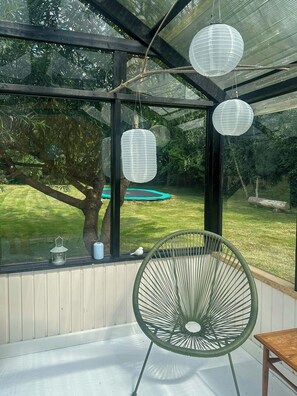 Une véranda avec un fauteuil vert, des lanternes en papier suspendues et une vue sur une pelouse et un arbre.