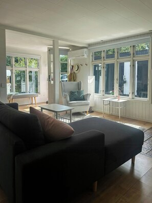 A modern living room with a sofa, armchair, and a small table. There are large windows with curtains, a ceiling fan, and a hanging plant. - Summer cottage with orangery and garden, Falster (Væggerløse)