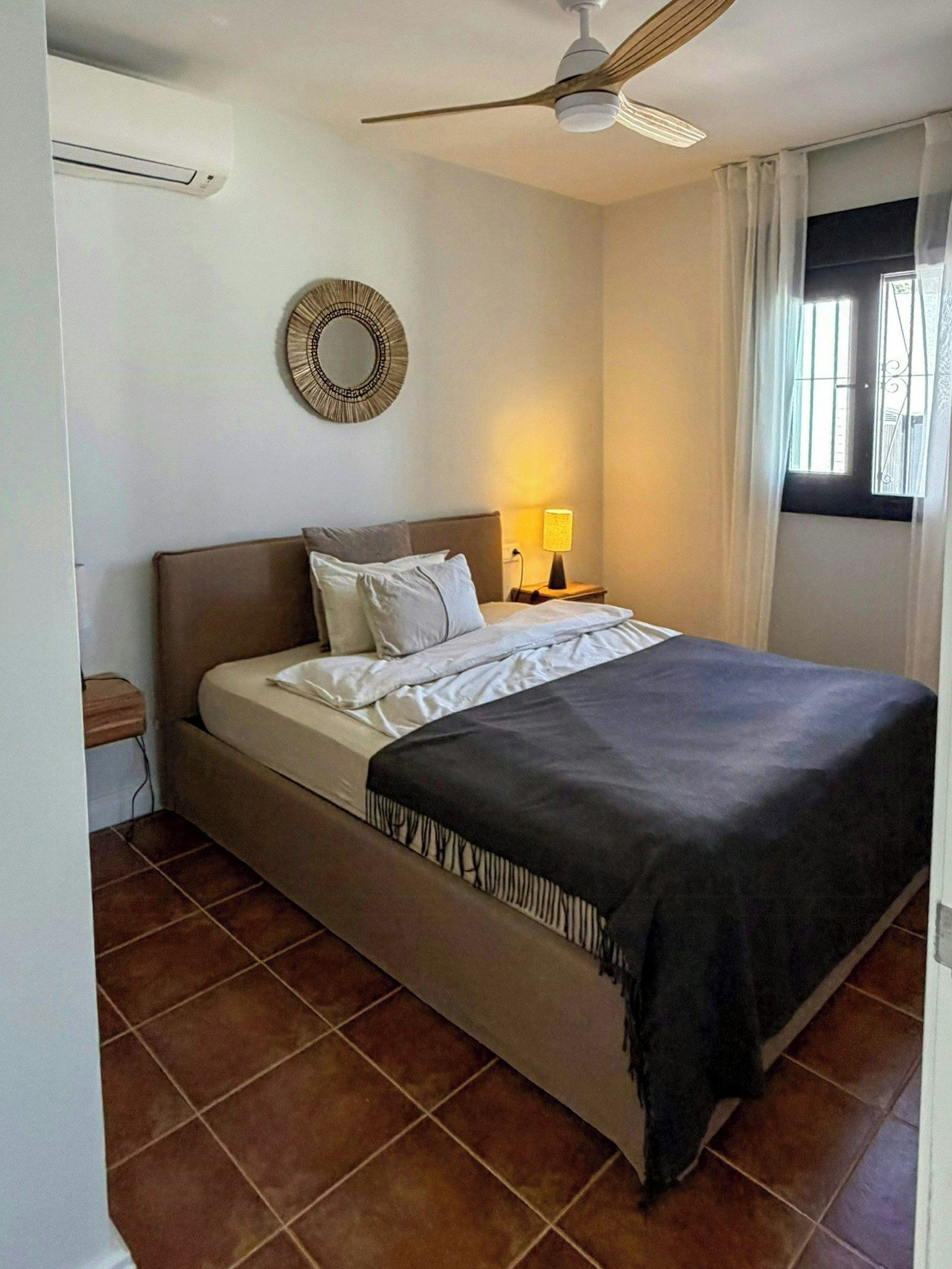 A bedroom with a bed, a nightstand, a lamp, a ceiling fan, a window with curtains, and a wall-mounted mirror.