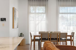 Ein modernes Wohnzimmer mit Essbereich, ausgestattet mit einer Sofagarnitur, Holzstühlen, einem Couchtisch und einem großen Fenster mit transparenten Vorhängen.