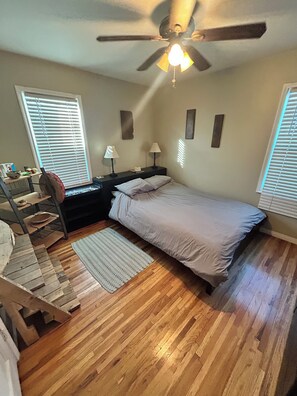 Ein Schlafzimmer mit einem Bett, Deckenventilator, Nachttisch, Lampe, einem Fenster mit Jalousien und Holzboden.