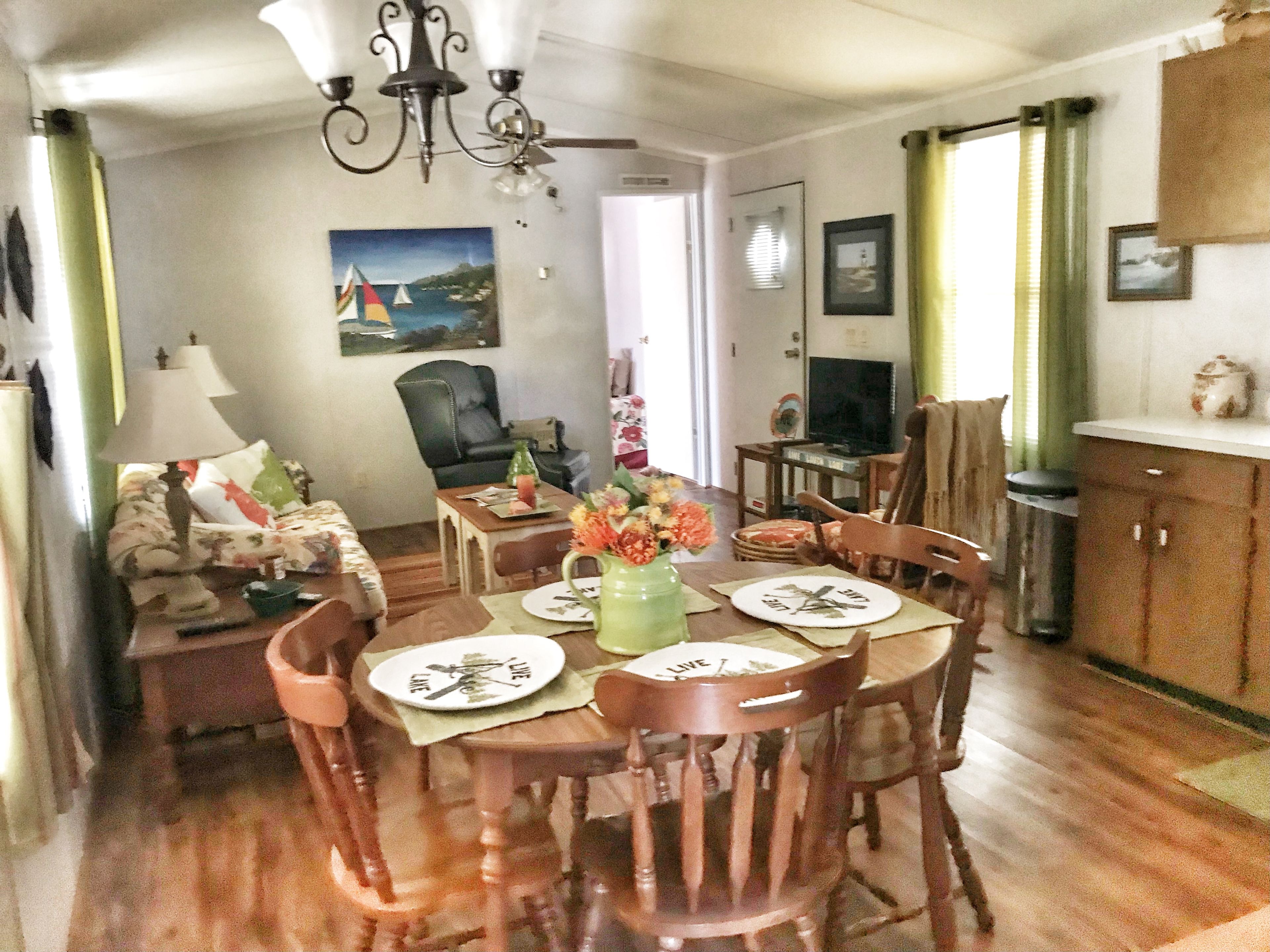 Un coin repas avec une table dressée pour quatre personnes, un lustre, un tableau au mur et un téléviseur en arrière-plan.