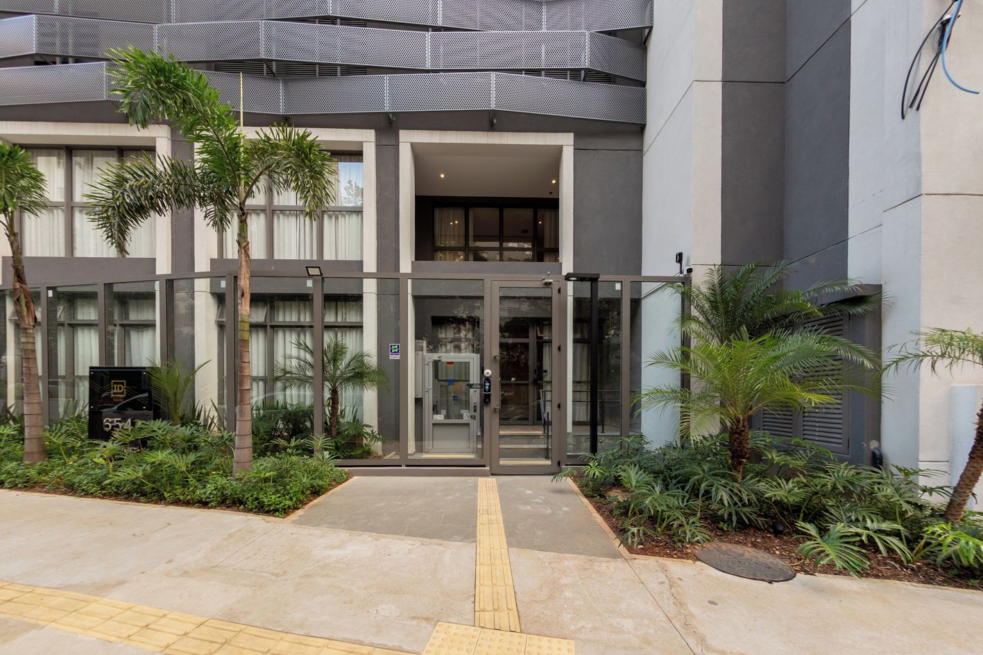 A modern building entrance with glass doors, a security keypad, and a small garden with palm trees.