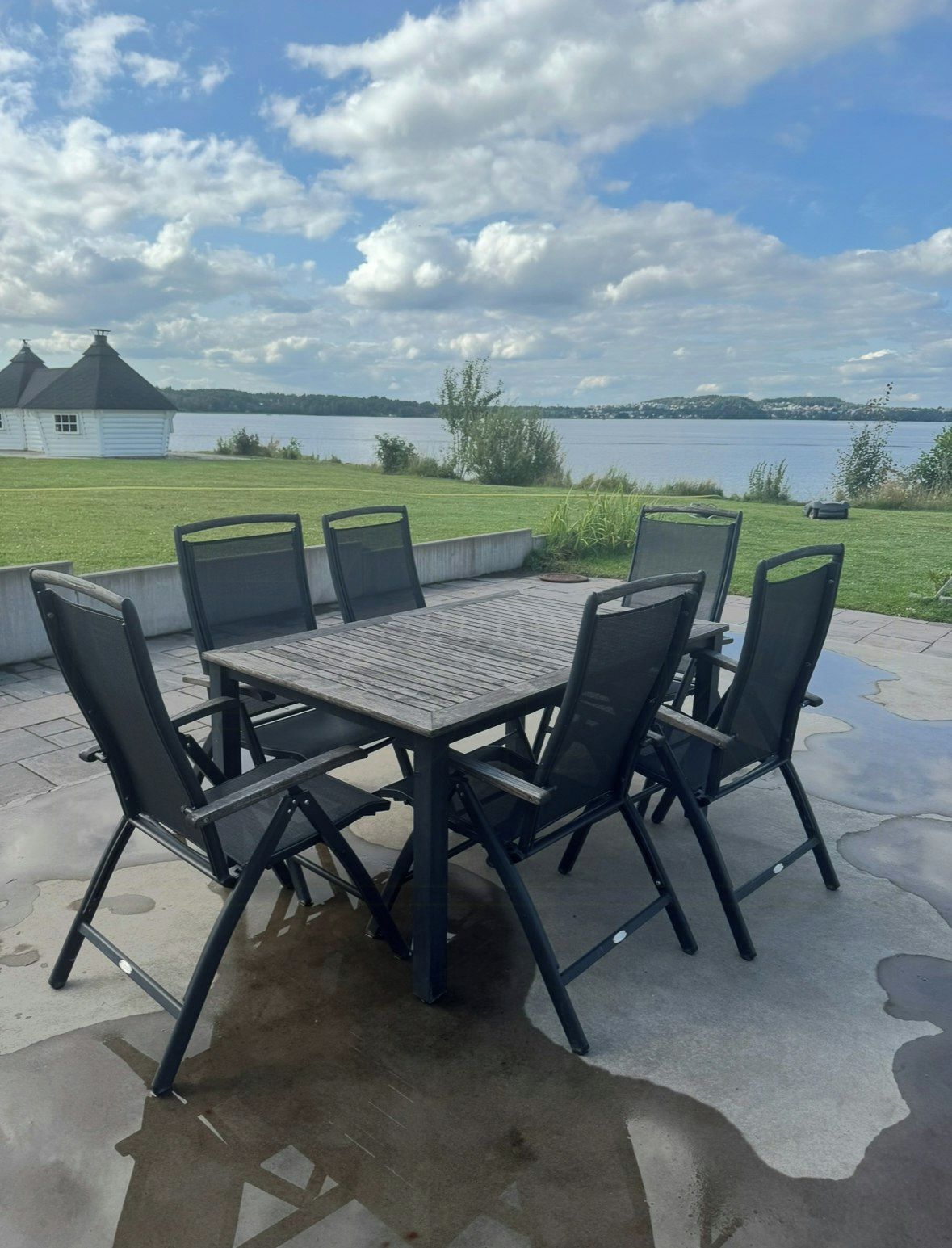 An outdoor patio with a table and chairs, a view of a lake, and a white building in the background.