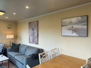 A living room with a sofa, a coffee table, a lamp, a painting, and a dining table with chairs.