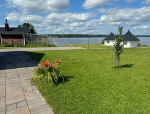 A lakeside property with a paved walkway, a grassy lawn, and a small white building with a conical roof.