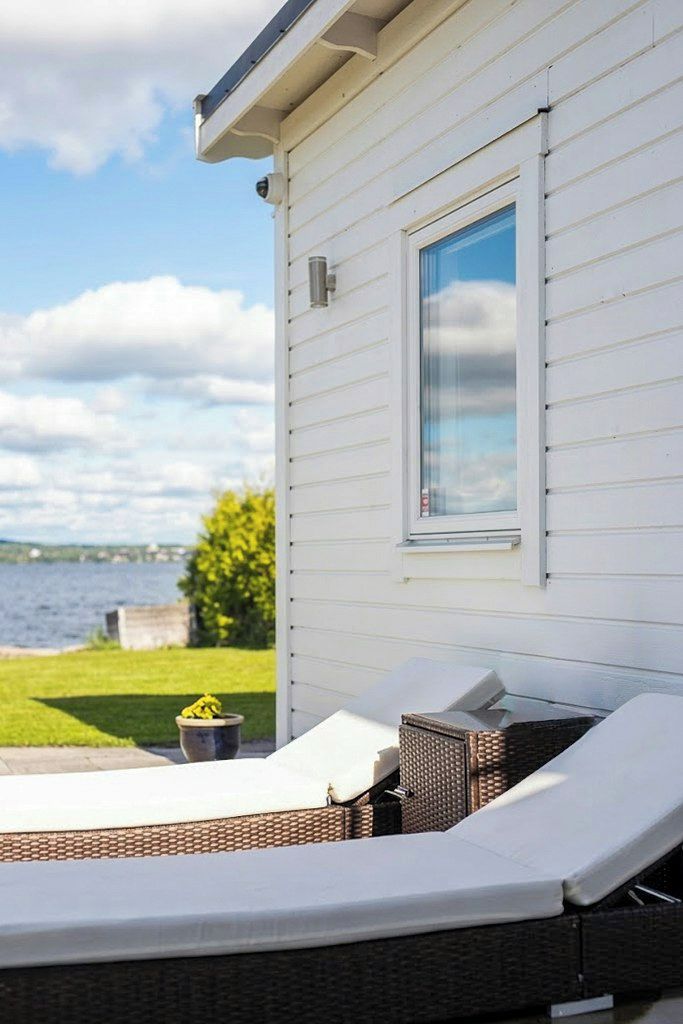 En vit byggnad med ett fönster som vetter ut mot en vattenyta. Där finns en loungehörna med en vit kudde och en flätstol.