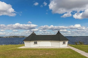 A white log cabin with a black roof, situated on a grassy area with a view of a body of water and a distant shoreline.
