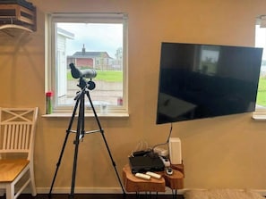 A room with a telescope on a tripod, a flat-screen TV mounted on the wall, and a small table with electronic devices.