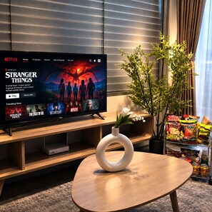 A modern living room with a flat-screen TV displaying a Netflix interface, a wooden coffee table, a potted plant, and snacks on a shelf. - Man Seaside Homestay - The Song Vung Tau (Thanh pho Ho Chi Minh)