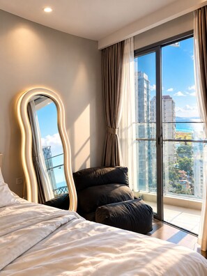 A bedroom with a large bed, a mirror, and a view of a cityscape through a balcony. - Man Seaside Homestay - The Song Vung Tau (Thanh pho Ho Chi Minh)