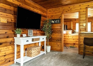 Interior de una cabaña de madera con un televisor en la pared, una mesa consola blanca con cajones, una planta en maceta y un letrero que dice 'HOME'.