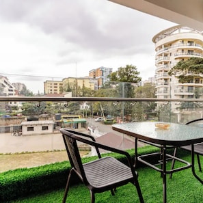 Een balkon met een glazen tafel en stoelen, met uitzicht op een straat met gebouwen en groen.