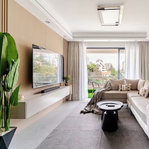 Een moderne woonkamer met een flatscreen televisie aan een witte console, een beige bank met decoratieve kussens, een ronde salontafel en een grote potplant.