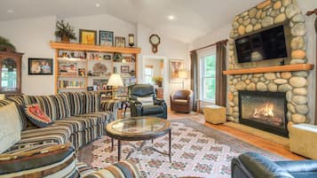 A cozy living room with a stone fireplace, a comfortable sofa set, a television mounted above the fireplace, and a bookshelf filled with books and decorative items.