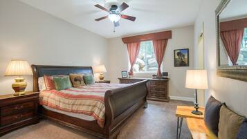 A bedroom with a bed, two bedside tables, a nightstand, a dresser, a mirror, a ceiling fan, and a window with curtains.
