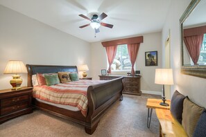 A bedroom with a bed, two bedside tables, a nightstand, a dresser, a mirror, a ceiling fan, and a window with curtains.
