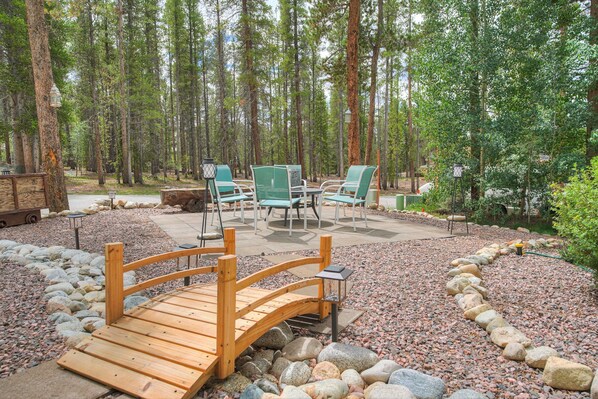 A wooden bridge leading to a seating area with chairs and tables, surrounded by a forest.