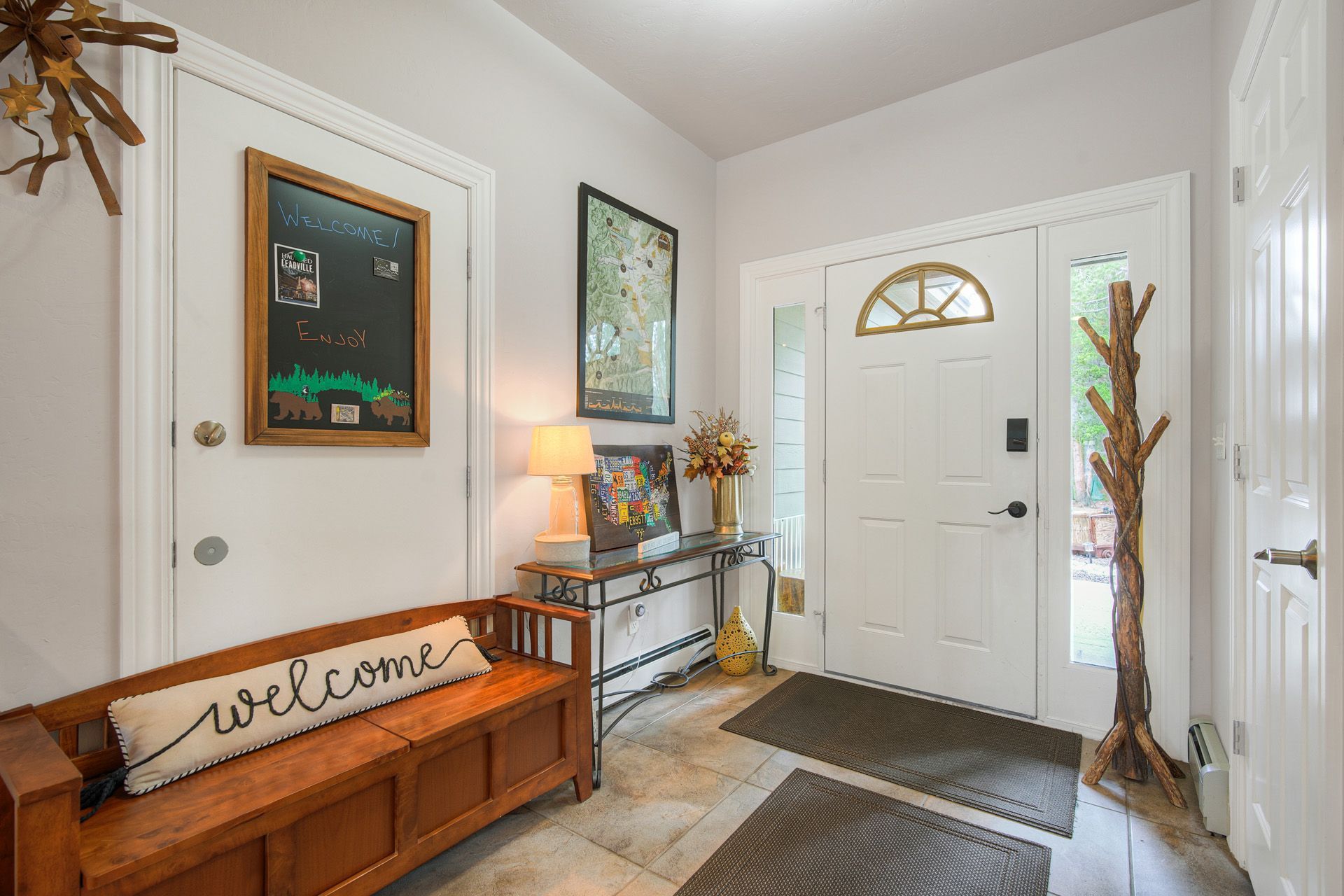 A wooden bench with a 'welcome' pillow, a framed map, a lamp, and a door with a decorative arch.