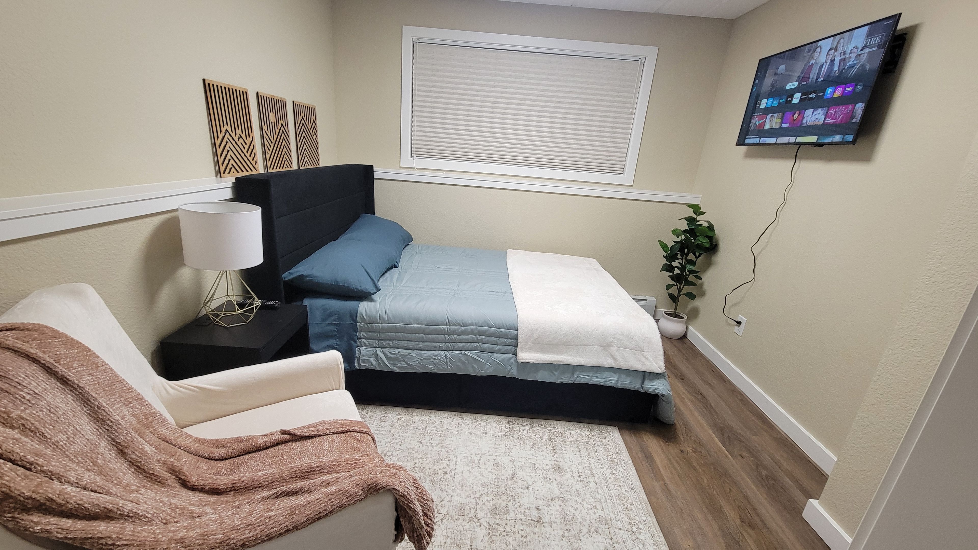A bedroom with a bed, a chair, a bedside table, a lamp, a TV mounted on the wall, and a window with blinds.