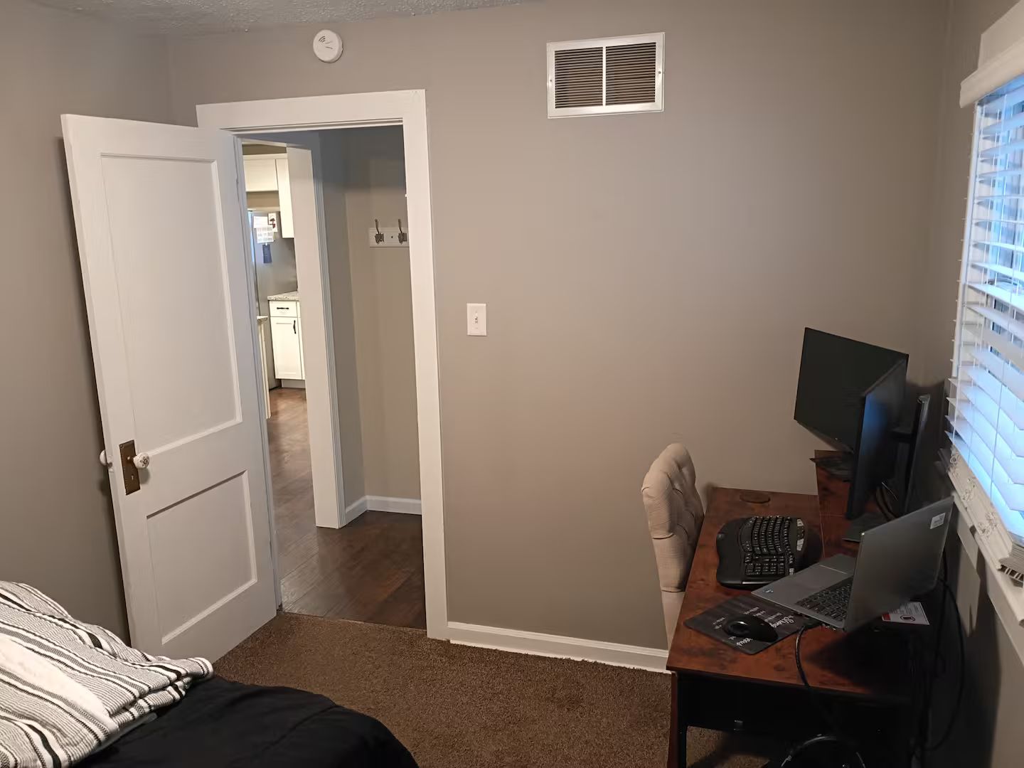 Ein Schlafzimmer mit einem Bett, einem Schreibtisch mit Computer, einem Fernseher und einem Fenster mit Jalousien.
