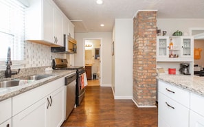 Cocina con armarios blancos, encimera de granito, pared decorativa de ladrillo y vistas al baño contiguo.
