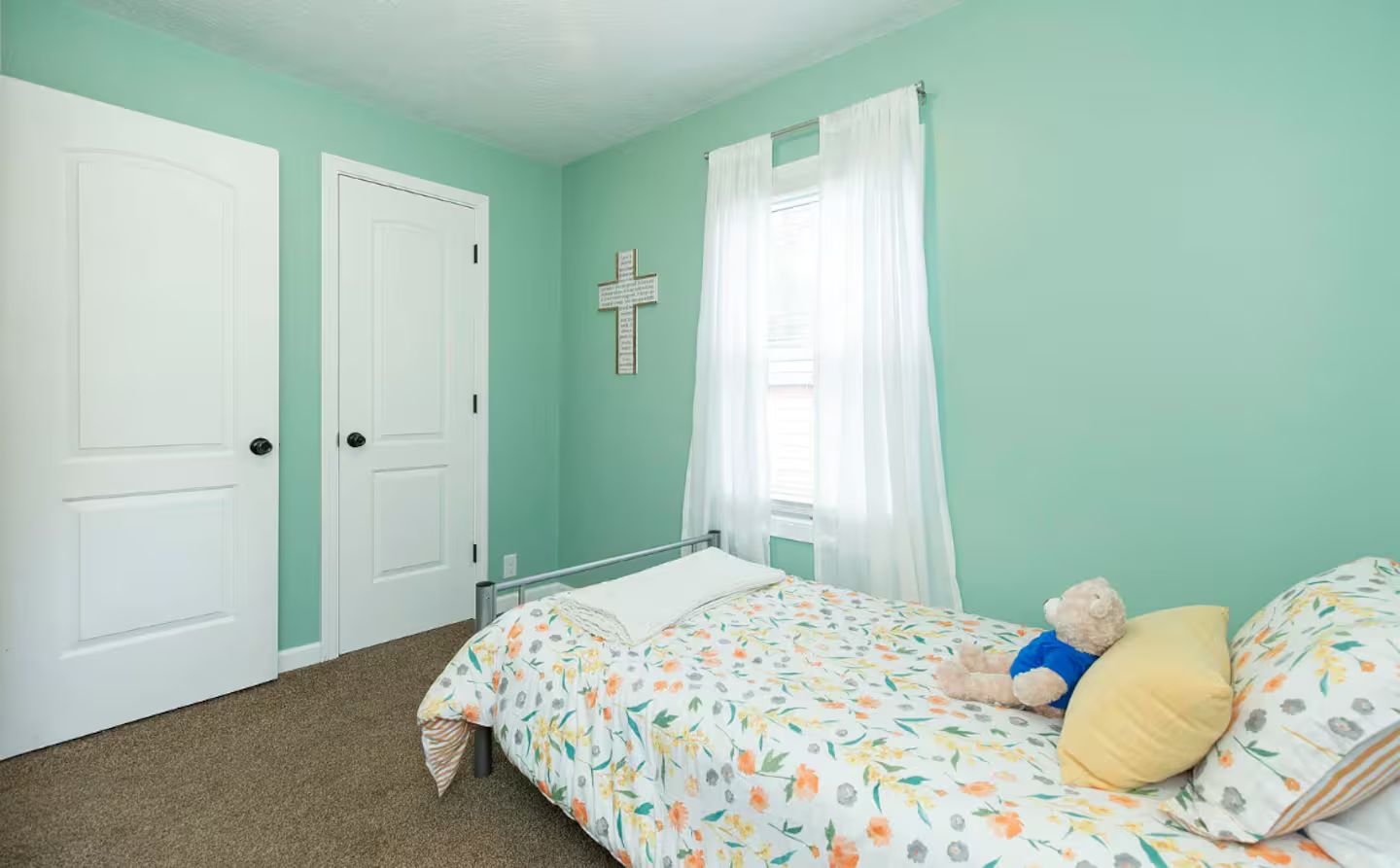 A bedroom with a bed, a teddy bear, and a cross on the wall.