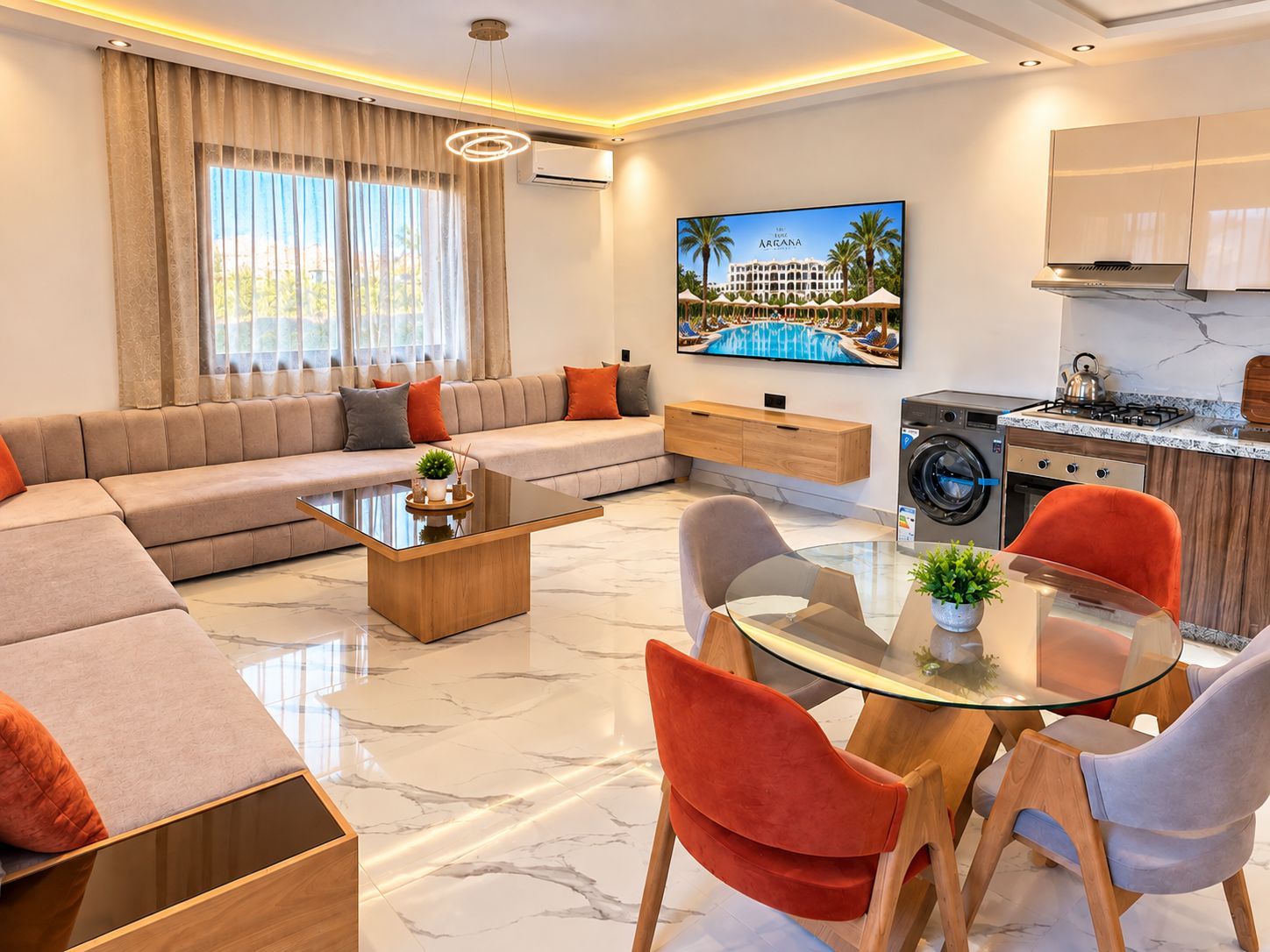 Una sala moderna con sofá, mesa de centro y sillas, un área de cocina con estufa y lavadora, y un televisor que muestra un resort.